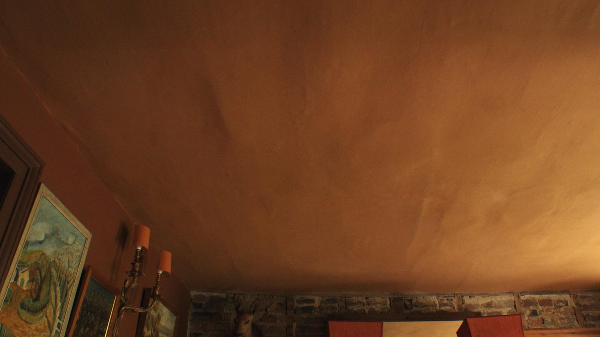 A warm, earthy orange ceiling with a softly textured, hand-finished surface above a room with rustic brickwork and framed artwork