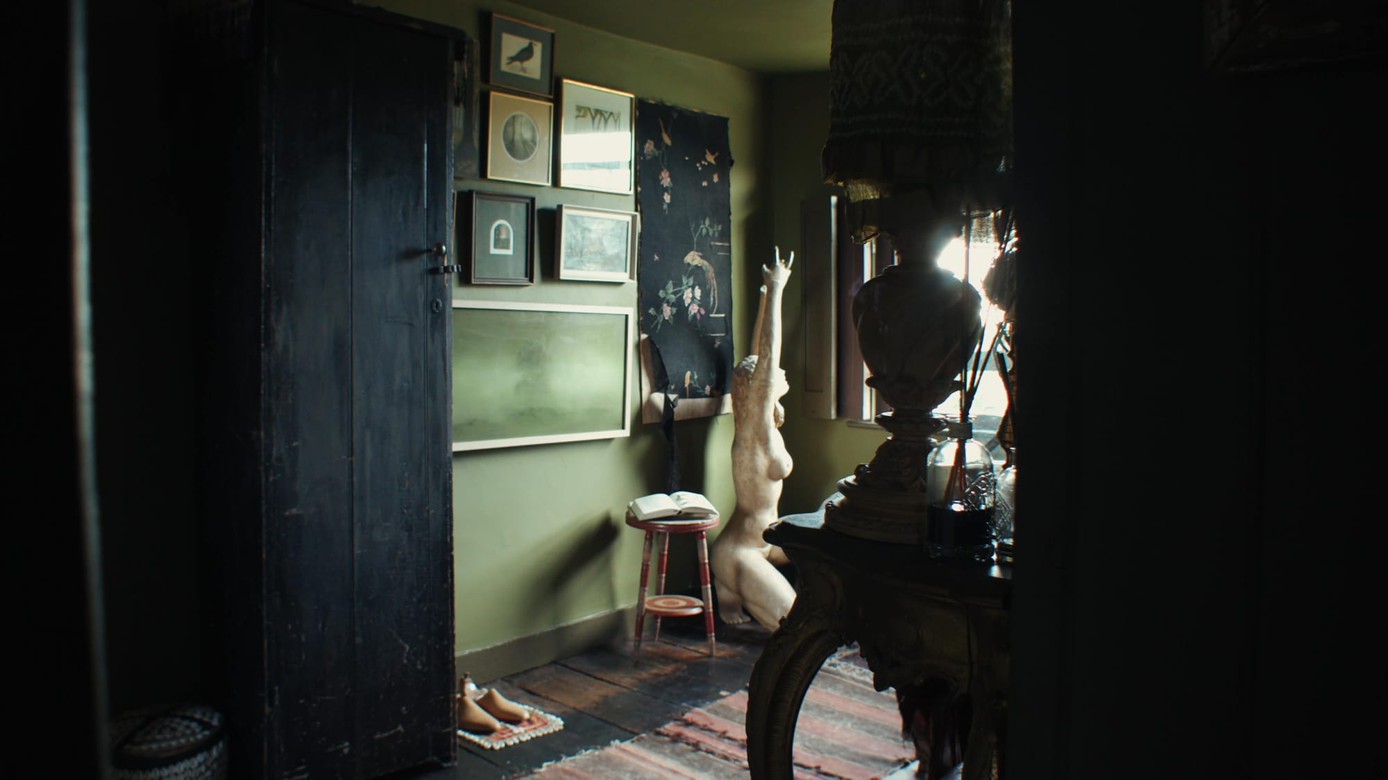 A dim, eclectic room with olive-green walls, a gallery of framed art, a distressed black wardrobe, a kneeling sculpted female figure with arms raised, and a small stool with an open book beside it
