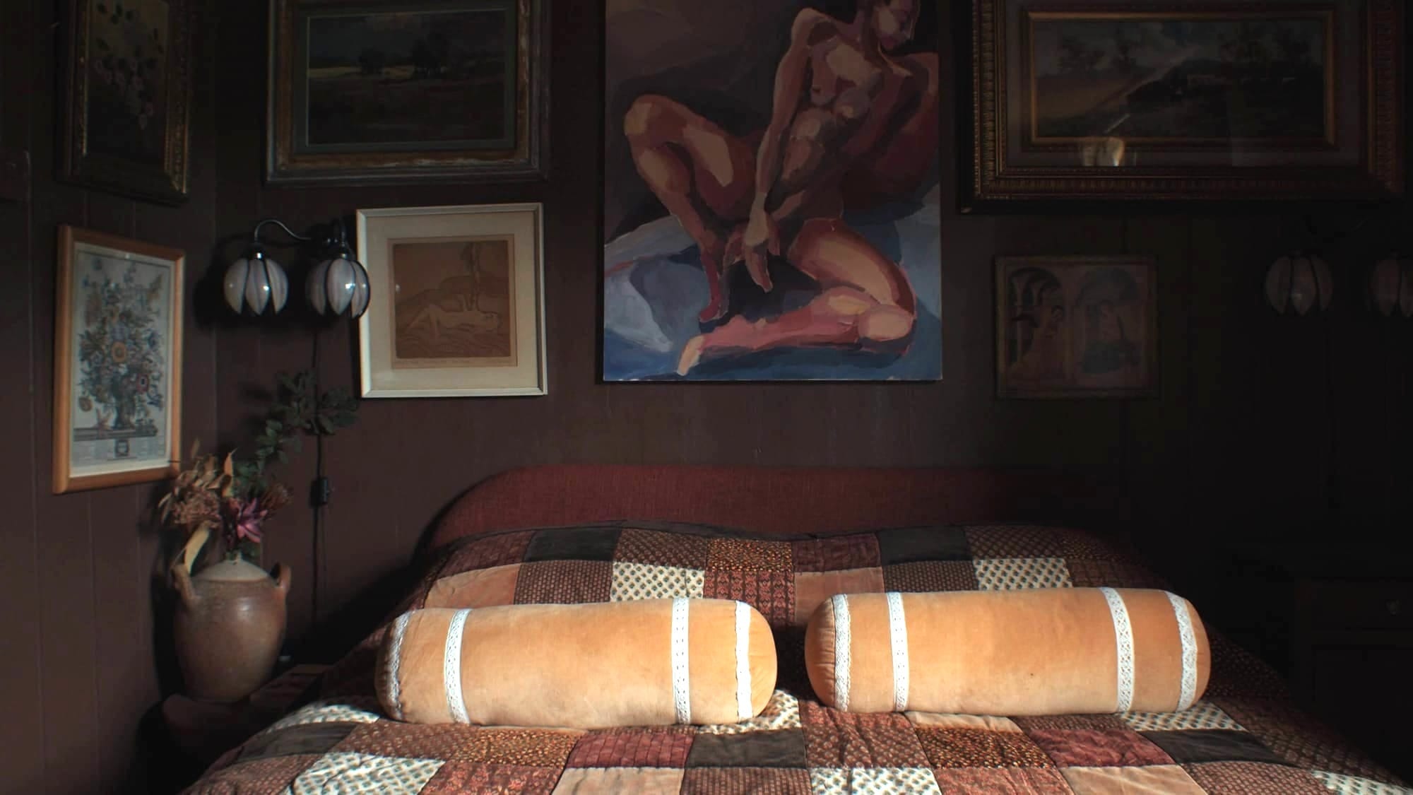 A dark, cosy bedroom with a patchwork quilt, two long cylindrical cushions, and a wall filled with framed artwork, including a large painting of a seated nude figure above the bed