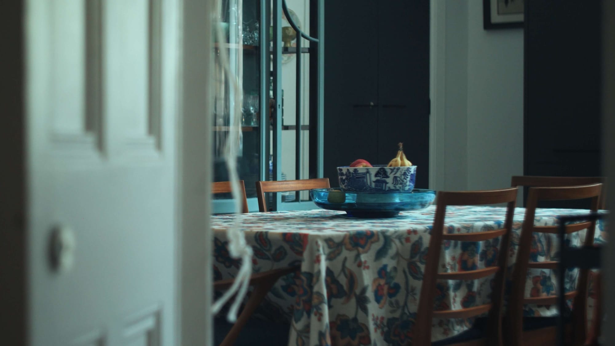 Dining table with a patterned tablecloth, wooden chairs, and a blue fruit bowl in a bright tenement kitchen