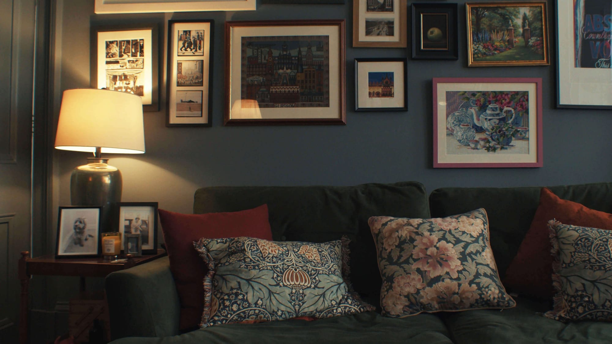 Gallery wall above a green velvet sofa with patterned cushions, warm lamp light, and framed art in a cosy living room