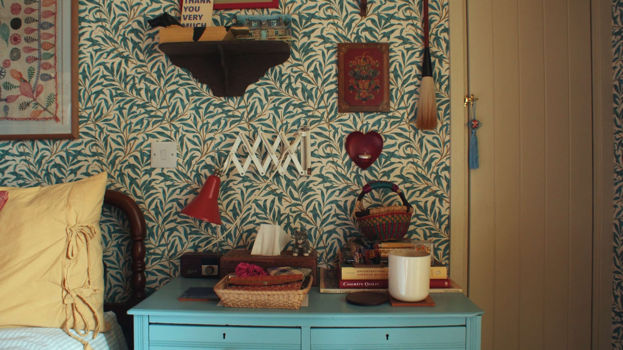 Bedside table against patterned wallpaper with books, baskets, wall lights and small decorative objects