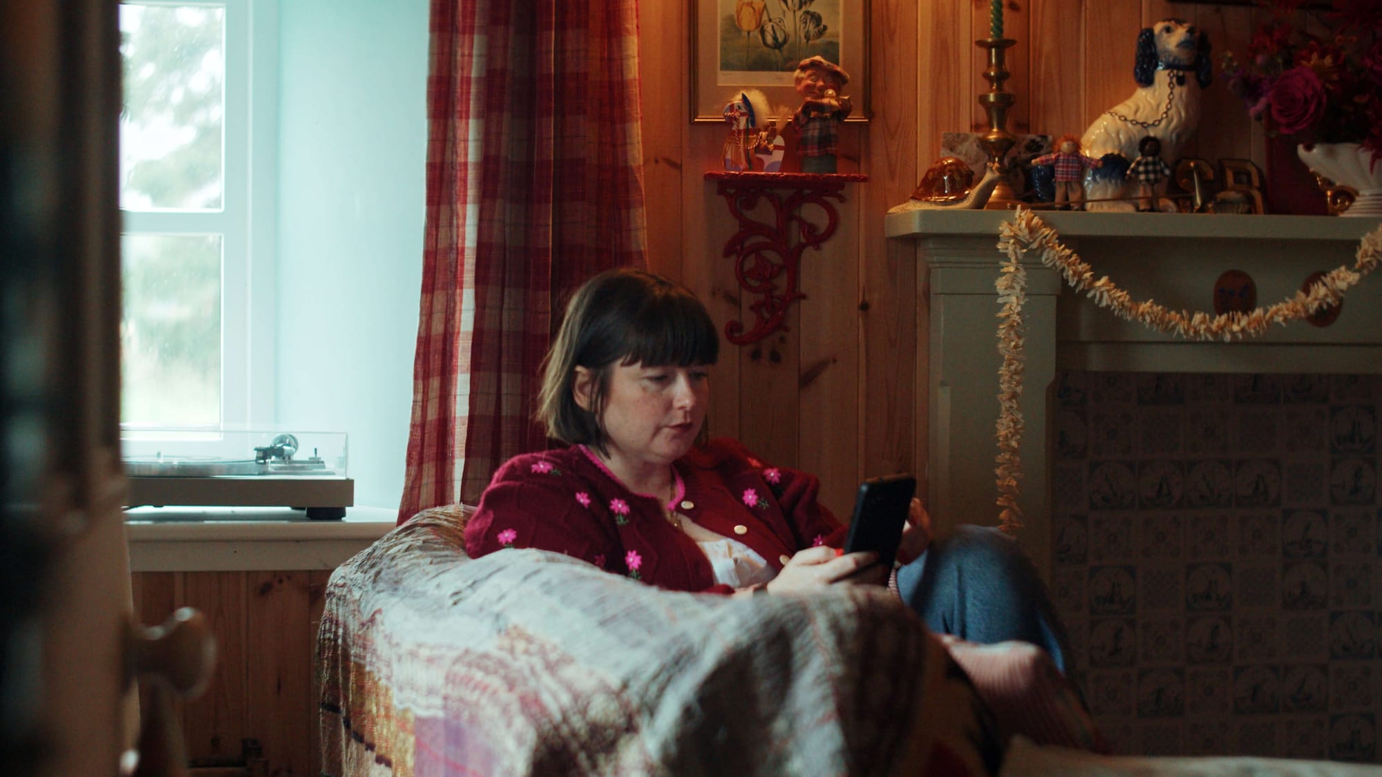 Rachel sitting in an armchair, looking at her phone beside a fireplace in a wood-panelled room