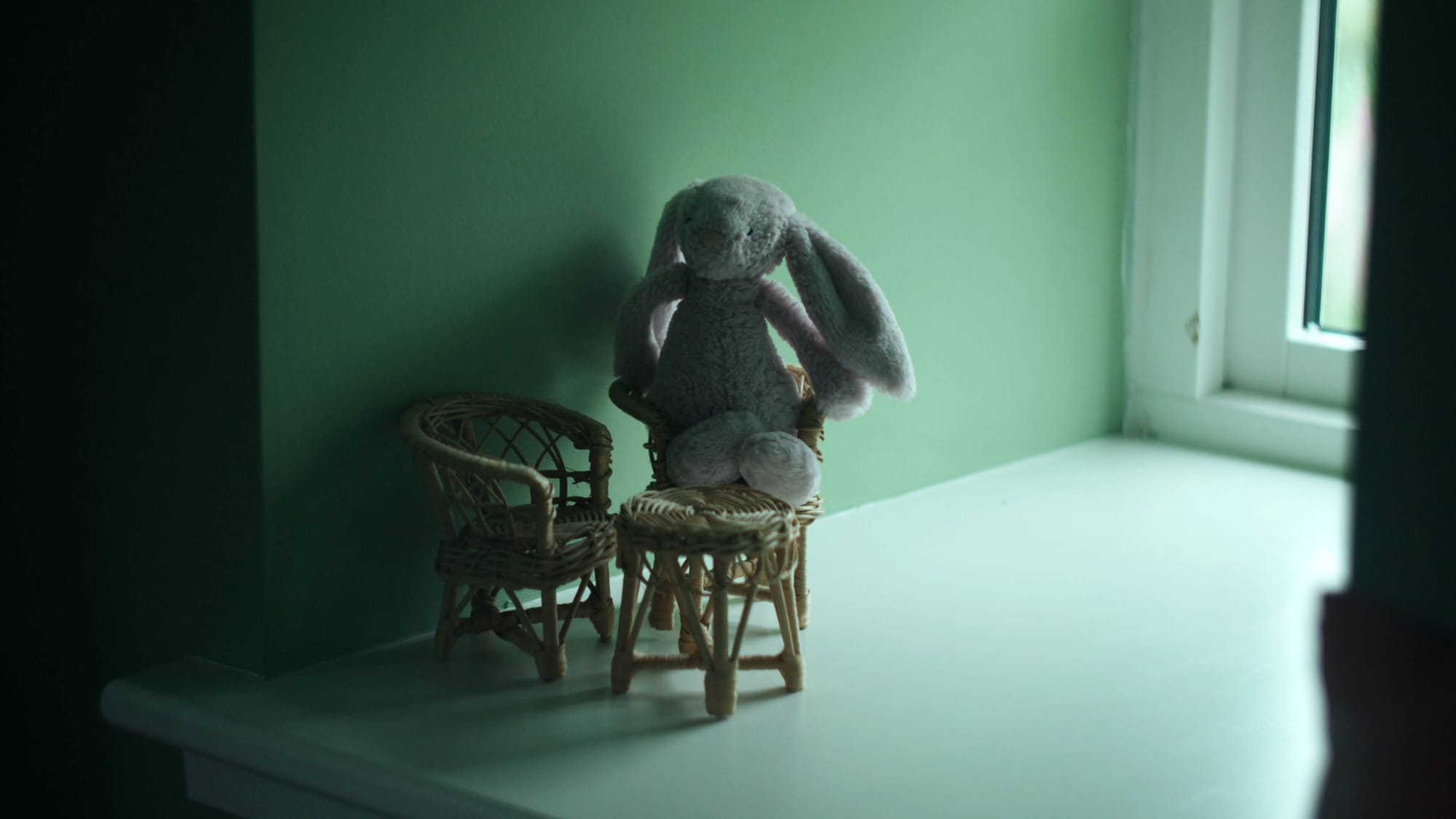 Soft toy rabbit sitting on small wicker chairs on a windowsill against a green wall