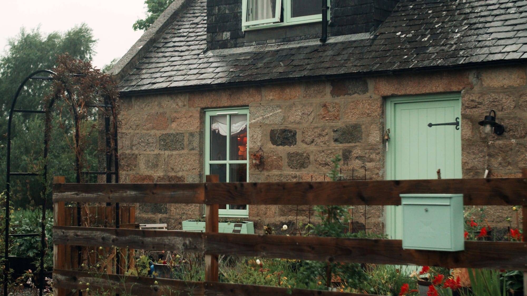 Stone cottage with pale green door, window and small garden behind a wooden fence