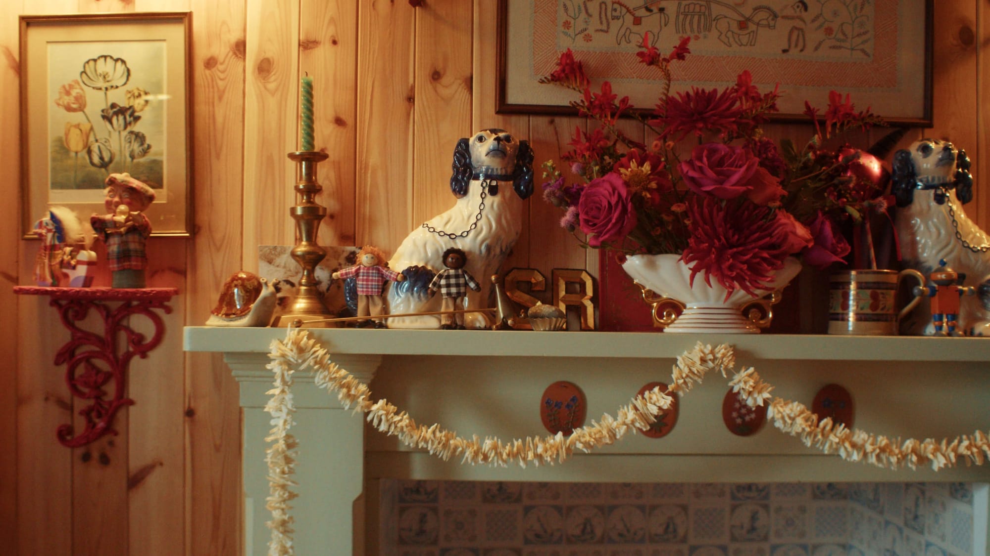Fireplace mantel with ceramic dog figures, candles, framed art and a vase of red flowers